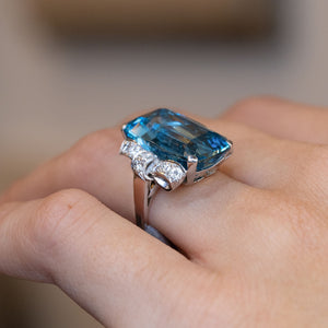 Side-angle view of the aquamarine ring worn on a hand, showcasing the raised platinum setting and decorative diamond shoulders.