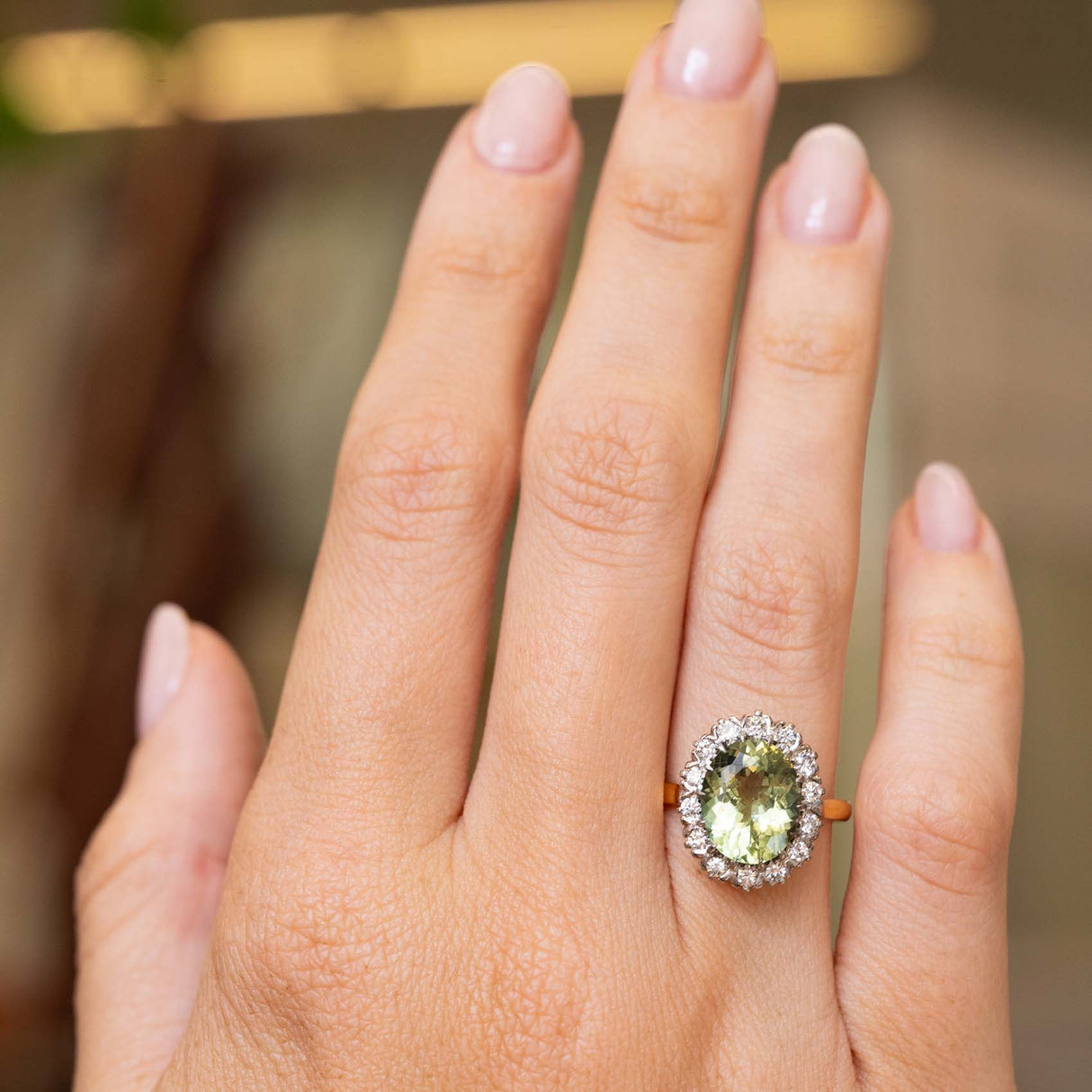 A close-up of a hand wearing a Vintage 1950s yellow gold cluster engagement ring set with an oval green beryl surrounded by a halo of diamonds with a yellow gold band.  Against a softly blurred interior background. 
