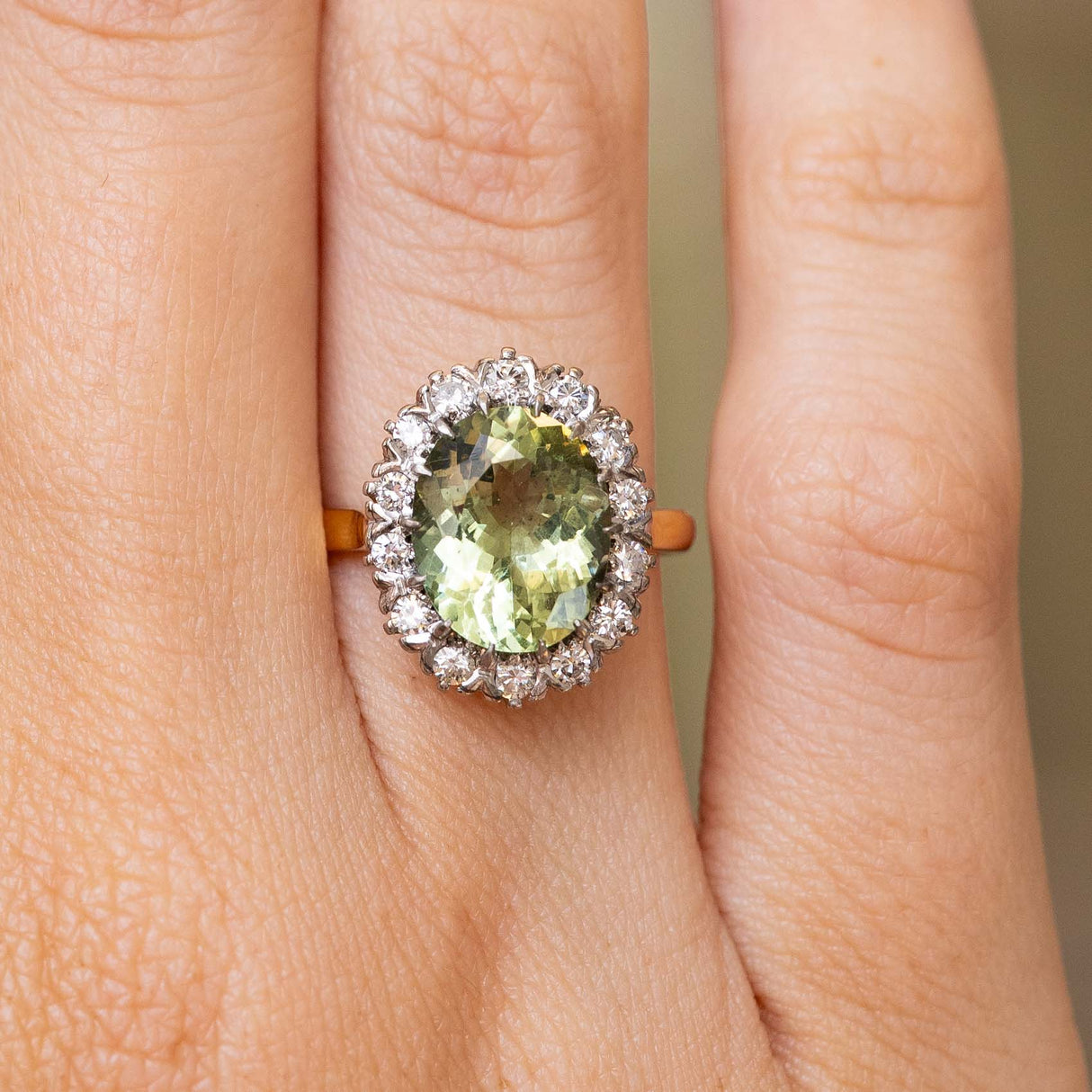 A close-up of a hand wearing a cluster engagement ring from the 1950s with a large oval light green beryl surrounded by a halo of diamonds and  set on a gold band.