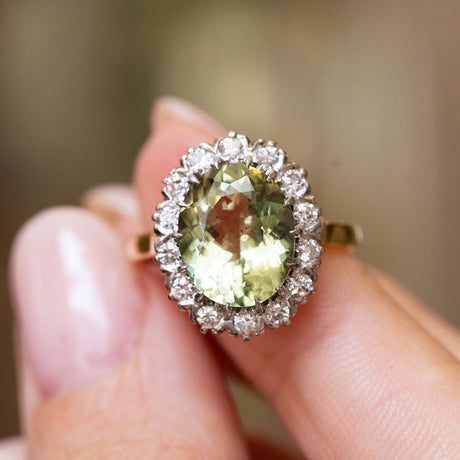 A close-up of an engagement ring being held between fingers. The Vintage 1950s ring features an oval-cut light green beryl surrounded by a halo of diamonds, set on a yellow gold band.