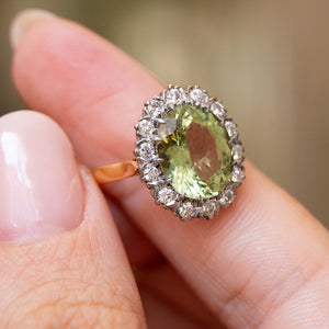 A close-up of a Vintage 1950s ring being held between fingers. The ring features a large oval light green beryl surrounded by a halo of diamonds, set on a yellow gold band.  