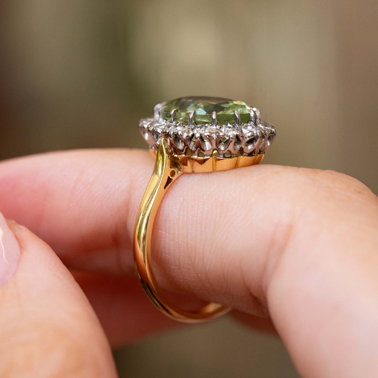 A close-up of a Vintage 1950s cluster engagement ring in yellow gold, showcasing the central light green beryl and surrounding cluster of diamonds. Worn on a person's finger. 