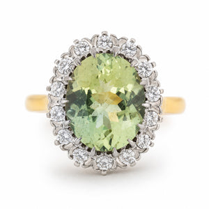 An Vintage 1950s cluster engagement ring, featuring a large light green beryl, surrounded by a halo of small clear diamonds, set on a yellow gold band against a white background.