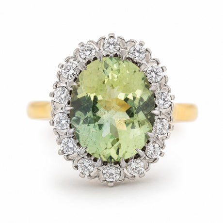 An Vintage 1950s cluster engagement ring, featuring a large light green beryl, surrounded by a halo of small clear diamonds, set on a yellow gold band against a white background.