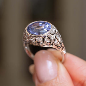 a detailed close image of the ornate details of an Antique, Belle Époque Ceylon colour-change sapphire and diamond cluster ring in platinum against a blurred background.