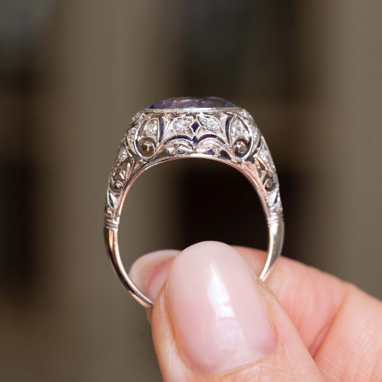 A platinum Antique, Belle Époque Ceylon colour-change sapphire and diamond cluster ring held between fingers and shown from the side to display the profile of the ring