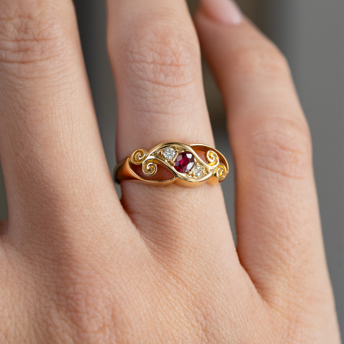 Close finger detail highlighting the rich red ruby, flanking diamonds and intertwined scrolling yellow gold motifs.