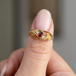 Handheld portrait of the ruby and diamond three-stone ring, displaying the decorative openwork and curled gold details.