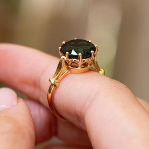A vintage 1980s cocktail ring with a yellow gold crown-like setting and a round dark green tourmaline, shown worn on a person's finger against a soft blurred background.