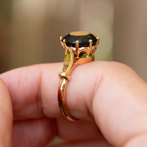 A close up of a Vintage, 1980's cocktail ring set with a dark green tourmaline in a yellow gold setting and band, shown being worn on a finger against a blurred warm background.