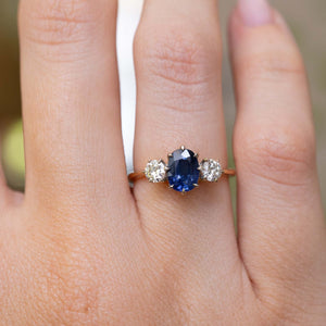 A close-up image of a hand wearing an Antique, Art Deco blue oval sapphire and diamond trilogy engagement ring with a yellow gold band worn on a finger against a blurry background.