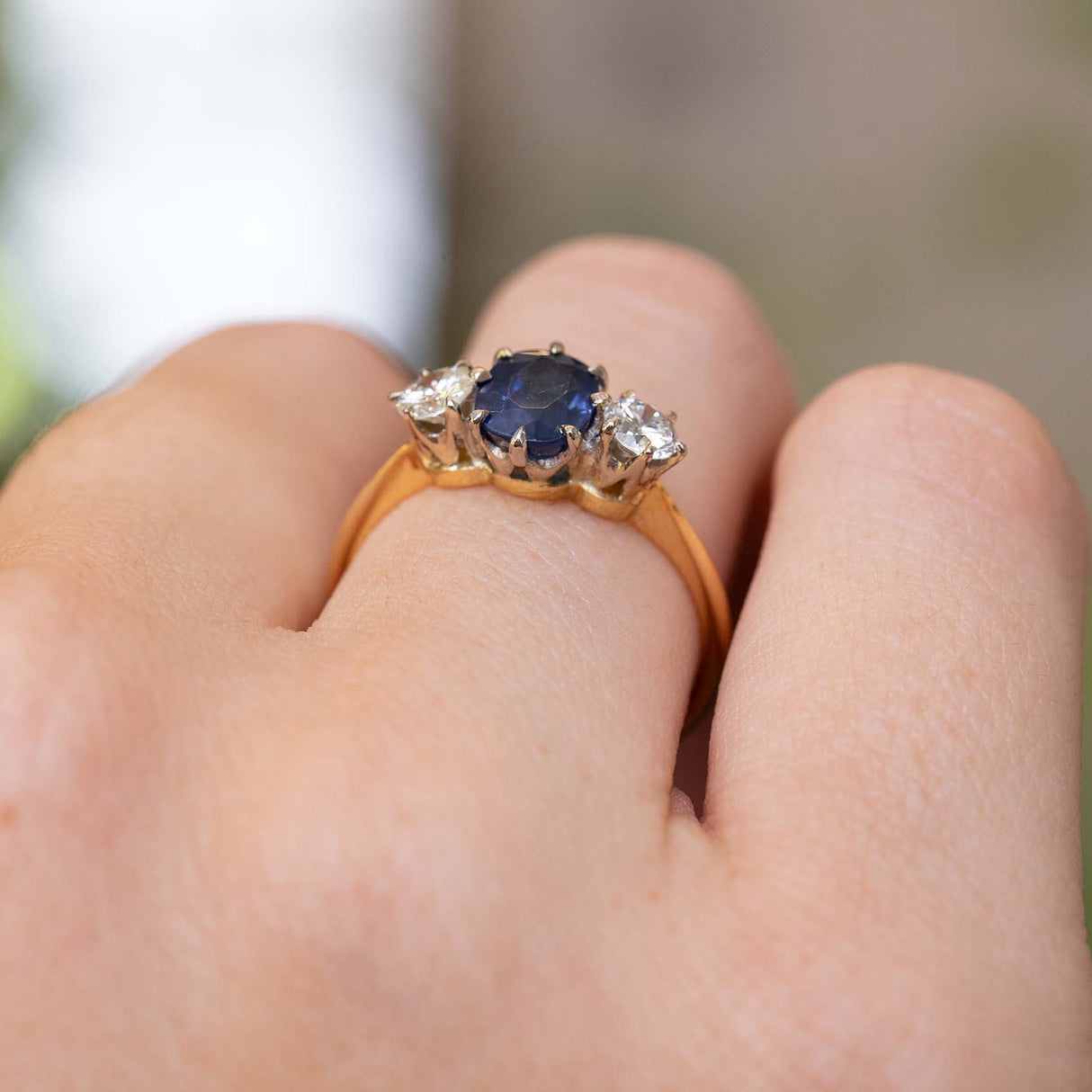 A close-up of an Antique, Art Deco sapphire and diamond three-stone engagement ring on a yellow gold band worn on a person's finger and seen from a low angle to show the setting prongs.