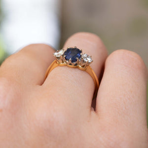 A close-up of an Antique, Art Deco sapphire and diamond three-stone engagement ring on a yellow gold band worn on a person's finger and seen from a low angle to show the setting prongs.