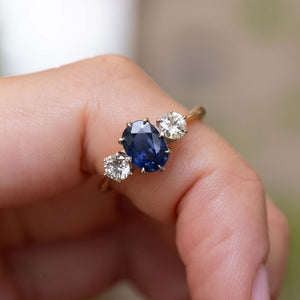 A close-up of a hand wearing a gold ring Antique, Art Deco blue oval-cut sapphire and diamond three-stone engagement ring on a yellow gold band, set against a softly blurred natural background.