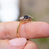 A close-up of an Antique, Art Deco blue sapphire and diamond three-stone engagement ring on a yellow gold band worn on a person's finger against a softly blurred background and showing off the prong setting style.