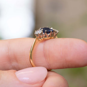 A close-up of an Antique, Art Deco blue sapphire and diamond three-stone engagement ring on a yellow gold band worn on a person's finger against a softly blurred background and showing off the prong setting style.