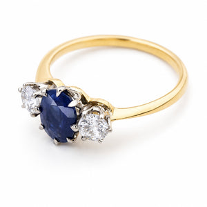 An image of a yellow gold Antique, Art Deco dark blue sapphire and diamond three-stone engagement ring on a white background and shown at a side angle to display the setting style.