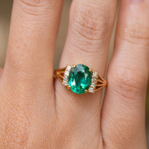 A close-up of the central emerald and flanking diamonds of a vintage 1970s engagement ring.  The ring is worn on a finger and showcases the yellow gold shoulder details. 