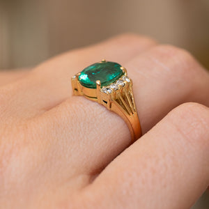 A close-up of a yellow gold engagement ring from the 1970s set with a large oval green emerald center stone and diamond accent stones, worn on a finger.