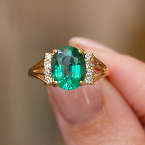 A yellow gold Vintage, 1970s emerald and diamond engagement ring featuring a large oval-cut green emerald, accented by small round diamonds on the shoulders, held between fingers against a soft beige background.