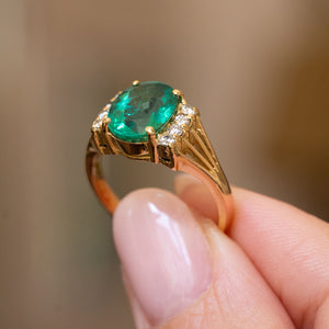 A close-up of a vintage yellow gold engagement ring from the 1970s set to centre with a large green emerald and flanked on either side with found diamonds.  Held between two fingers against a soft blurred beige background.