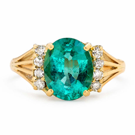A yellow gold Vintage, 1970s emerald and diamond engagement ring featuring a large central oval-cut emerald, accented by small clear round diamonds on each side of the band, set against a plain white background.