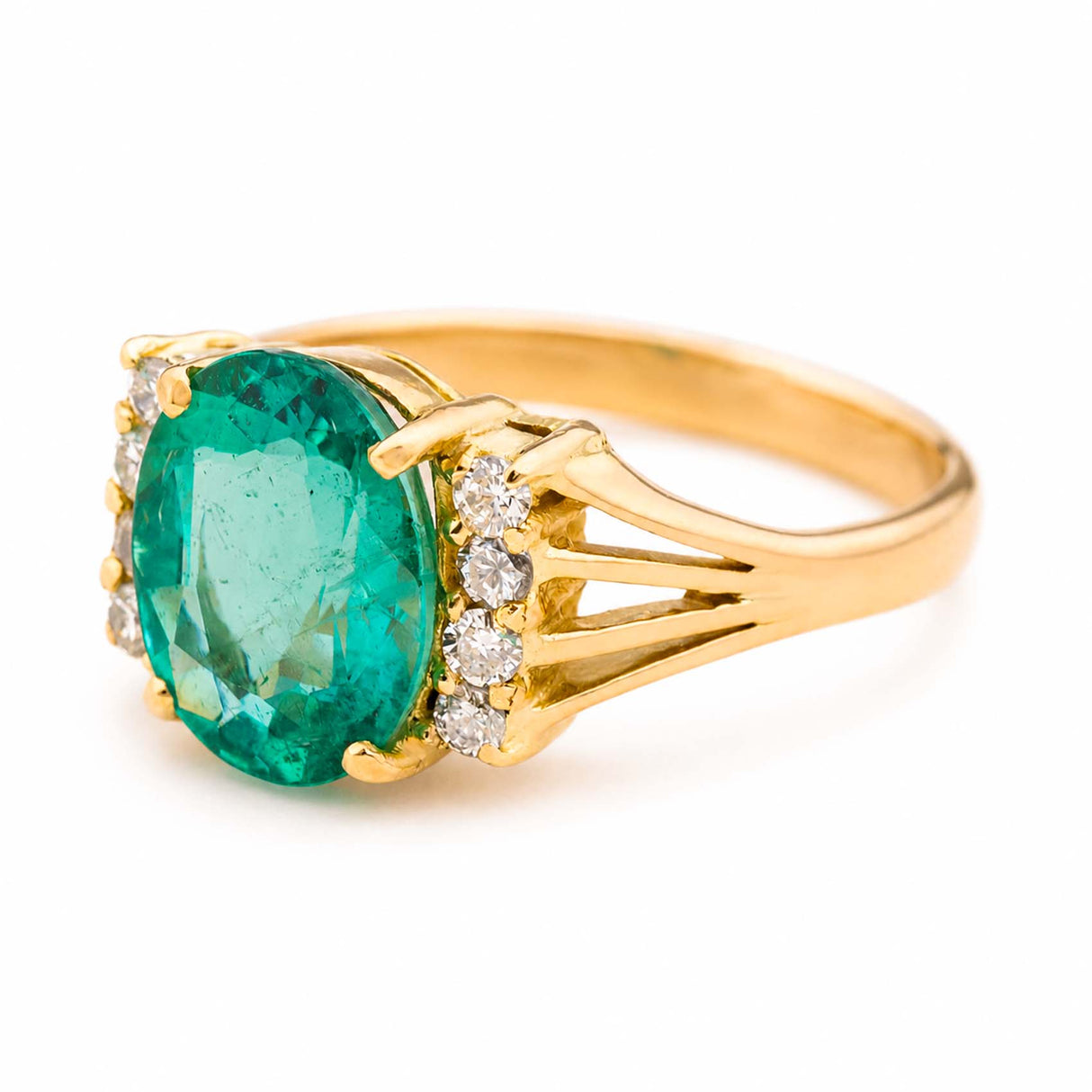 A yellow gold Vintage, 1970s emerald and diamond engagement ring shot from the side to showcase the central green emerald and the diamonds that flank it on either side.  Set against a plain white background.