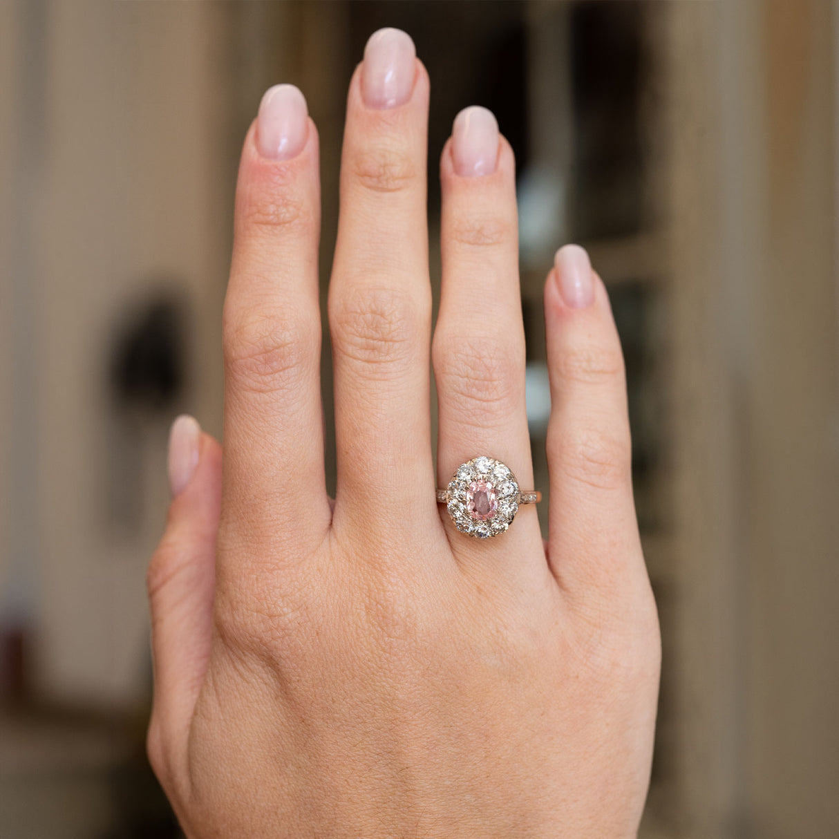 Vintage orange-pink sapphire and diamond cluster ring worn on the hand, featuring an oval orange-pink sapphire surrounded by old-cut diamonds.