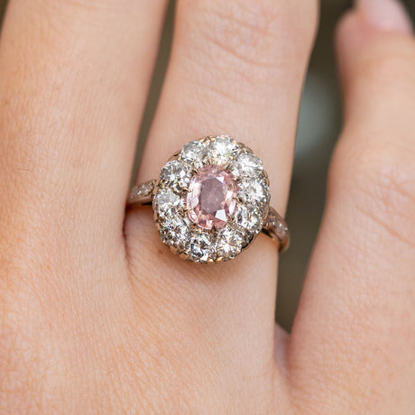 Vintage orange-pink sapphire and diamond cluster ring shown on the finger.