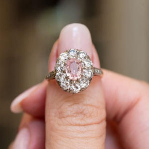 An oval orange-pink sapphire framed by a halo cluster old-cut diamonds worn on a thumb.