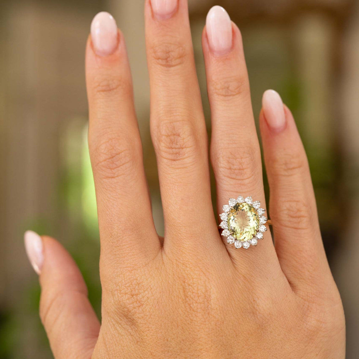  An outstretched hand wearing a vintage 1960s green beryl and diamond cluster engagement ring in yellow An outstretched hand wearing a vintage 1960s green barrel and diamond cluster engagement rang in yellow gold against a softly blurred background.