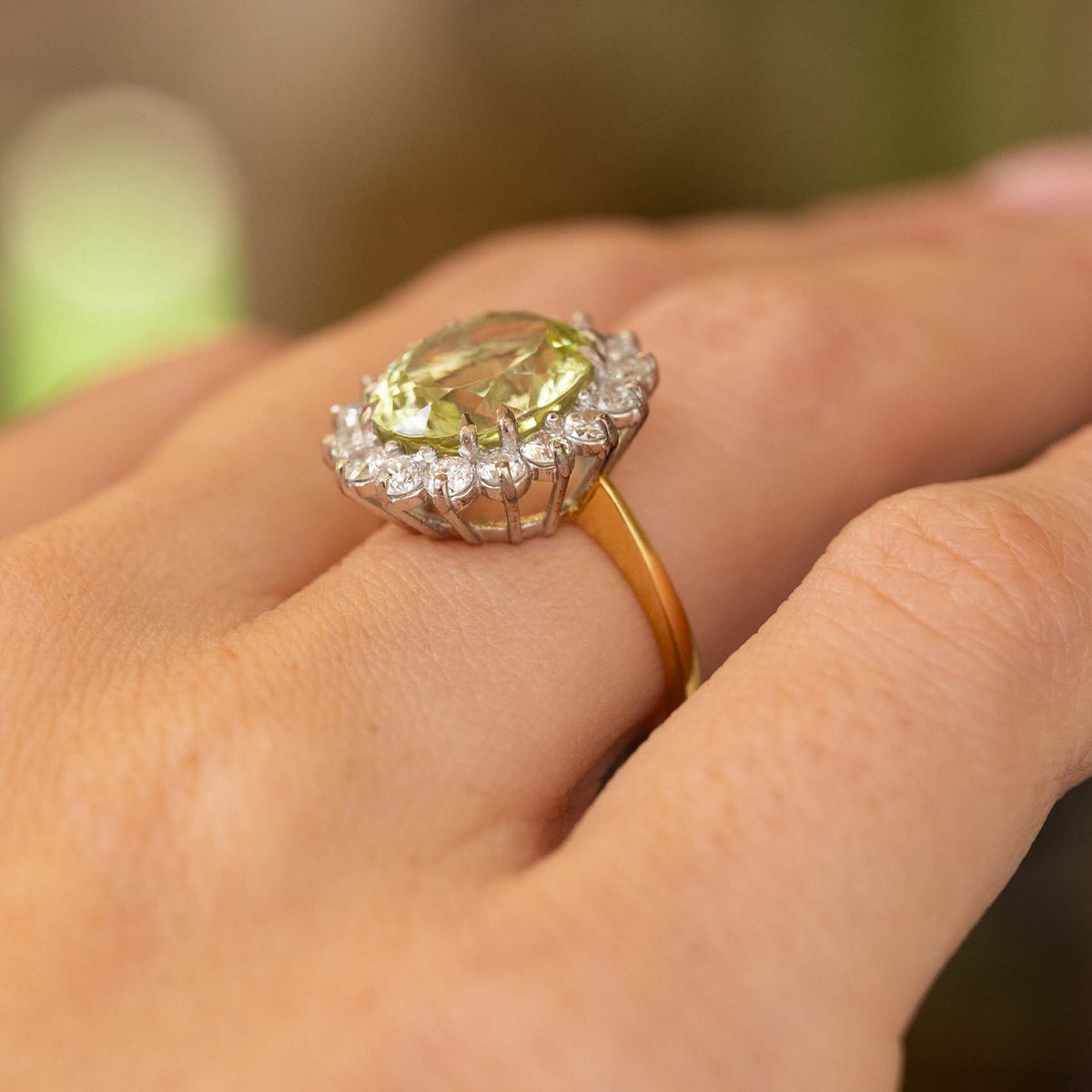 A close-up image of a yellow gold Vintage, 1960s Green beryl and diamond cluster engagement ring worn on the finger and pictured from a low angle to show off the basket style setting.