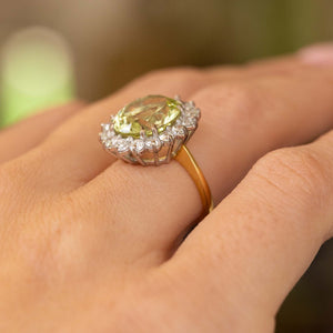 A close-up image of a yellow gold Vintage, 1960s Green beryl and diamond cluster engagement ring worn on the finger and pictured from a low angle to show off the basket style setting.