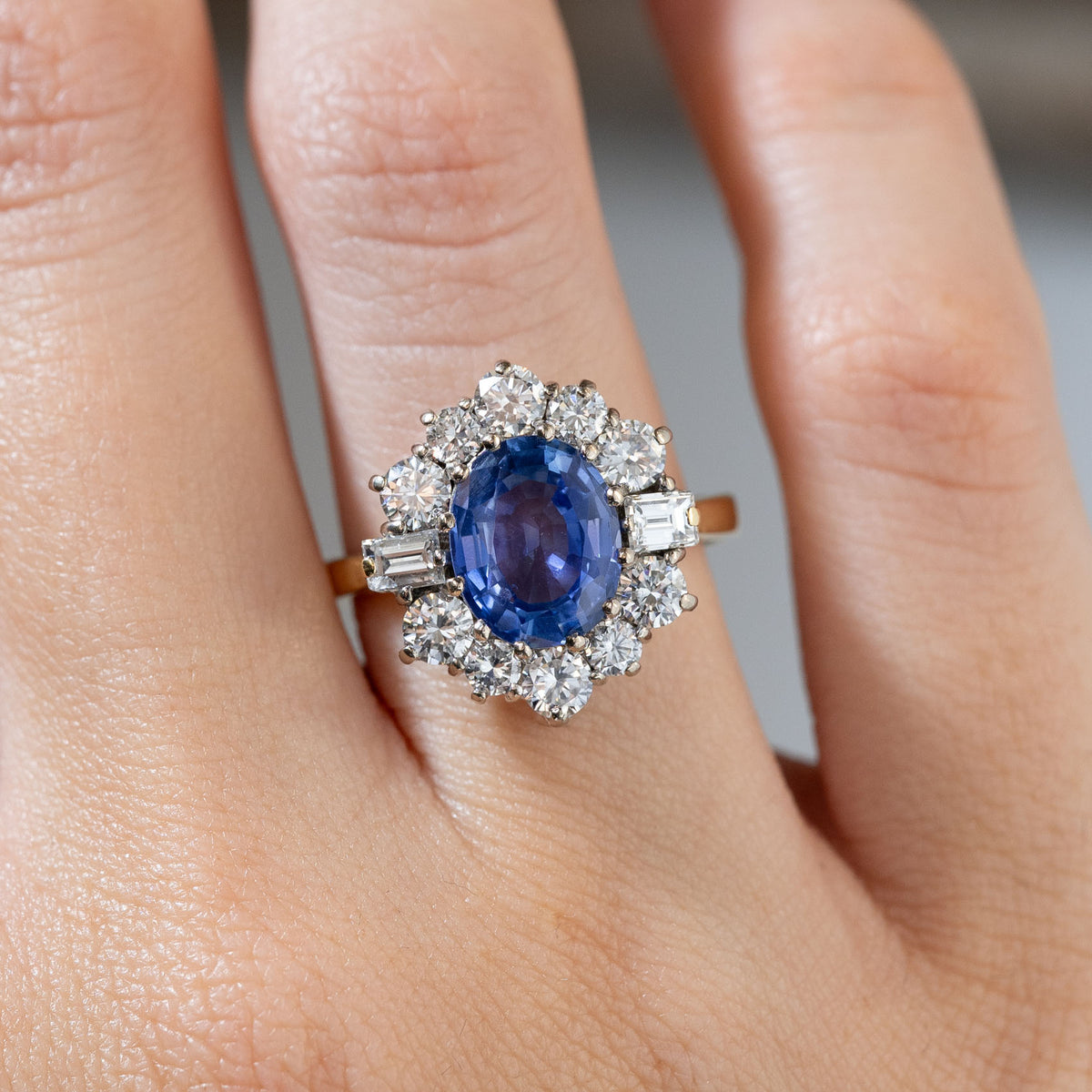 Close-up of a vintage 1980s blue sapphire and diamond cluster ring worn on a finger, featuring an oval blue sapphire framed by round brilliant-cut and baguette-cut diamonds.