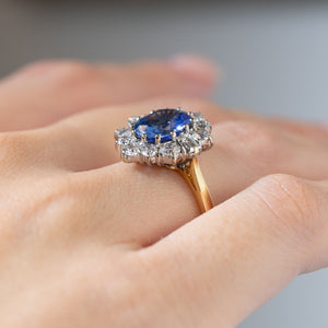 Side angle of a vintage 1980s blue sapphire and diamond cluster ring worn on a finger, revealing its raised platinum setting, oval sapphire and yellow gold shank.