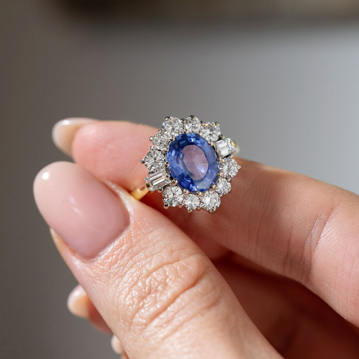 Detailed close-up of a vintage 1980s blue sapphire and diamond cluster ring held between fingers, highlighting the faceted oval sapphire and intricate diamond halo.