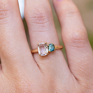 A close-up picture of a n Antique-inspired, colourless and blue Sapphire toi et moi two stone engagement ring in yellow gold worn on the finger against a blurred background.