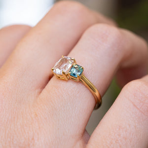 A close-up photograph of a hand wearing an antique-inspired, colourless and blue Sapphire toi et moi two stone yellow gold engagement ring seen from a slight angle to display the claw setting-style.