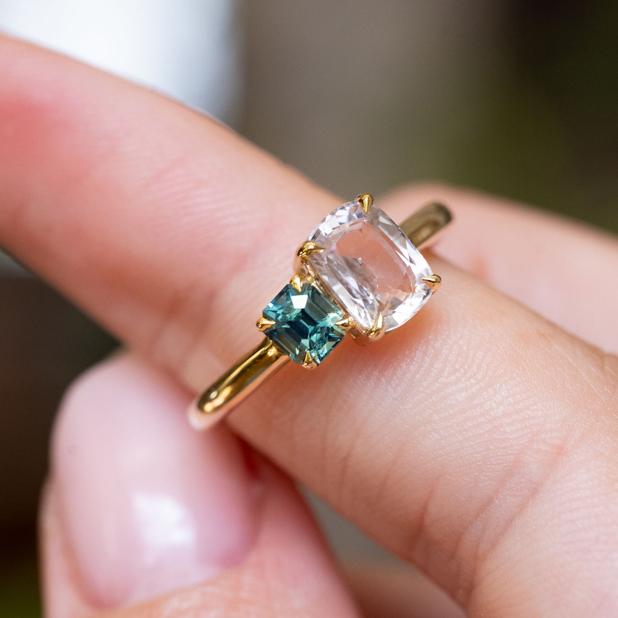 A close-up image of a gold antique-inspired engagement ring with a large colourless sapphire and a smaller teal-blue sapphire placed on a person's finger.