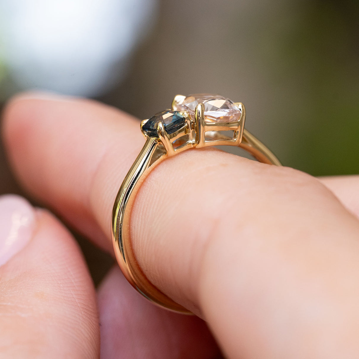 A detailed zoomed in image of a yellow gold Antique-inspired engagement ring featuring a colourless sapphire and a blue Sapphire, balanced on a finger and shown from the side profile to display the open-work gallery, against a softly blurred natural background.