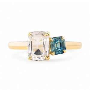 An antique-inspired yellow gold engagement ring featuring a large cushion-cut colourless sapphire and a smaller emerald-cut blue sapphire set on a plain white background.