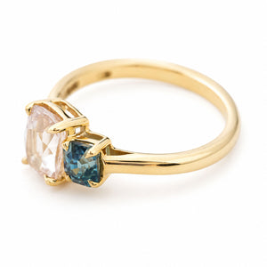 An antique-inspired yellow gold engagement ring featuring a large colourless sapphire and a smaller teal sapphire, set against a plain white background and photographed at an angle to show the details of the ring from the side. 