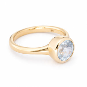 A close up of the side profile of a yellow gold antique inspired solitaire engagement ring, showcasing the rubber setting and white sapphire. Photographed against a clean white background. 