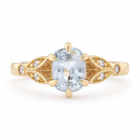 Vintage-inspired white sapphire and diamond engagement ring in yellow gold, featuring an oval-cut gemstone and decorative laurel leaf trefoil shoulders.