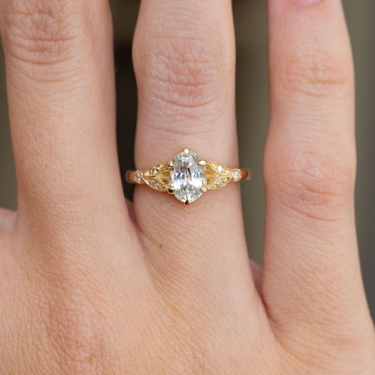 Close-up on-hand view of the oval white sapphire, framed by intricate diamond-set yellow gold shoulders.