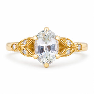Vintage-inspired white oval-cut sapphire and diamond engagement ring in yellow gold, with ornate openwork shoulders and fine beaded detailing.
