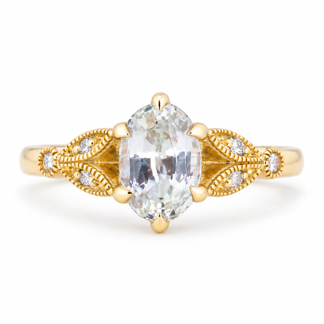 Vintage-inspired white oval-cut sapphire and diamond engagement ring in yellow gold, with ornate openwork shoulders and fine beaded detailing.