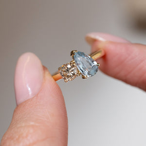 Hand-held close-up of the pale blue pear-cut sapphire and champagne-brown diamond in an antique-inspired yellow gold setting.