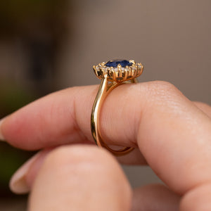 A close-up of a vintage inspired yellow gold cluster engagement ring, set with a blue sapphire and round-cut diamonds.  Displaying the side profile of the ring while worn on a finger and the basket and wedding fit design against a softly blurred neutral background.