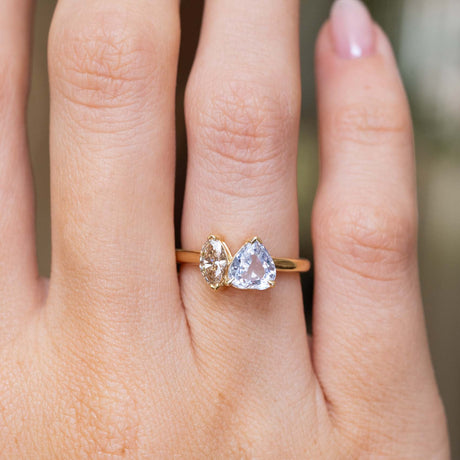 A close-up of a hand wearing an antique-inspired toi et moi two stone engagement ring of a colourless sapphire and a brown marquise-cut diamond on a yellow gold band to show the scale against a softly blurred green background.
